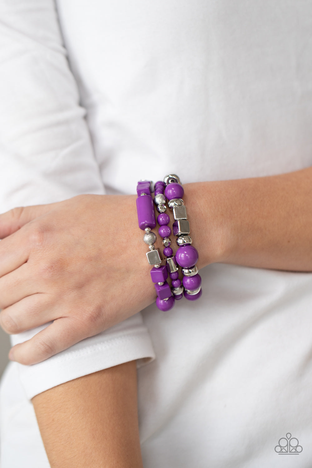 Featuring round, cube, and faceted shapes, a colorful collection of purple and silver beads are threaded along stretchy bands around the wrist, creating vivacious layers.