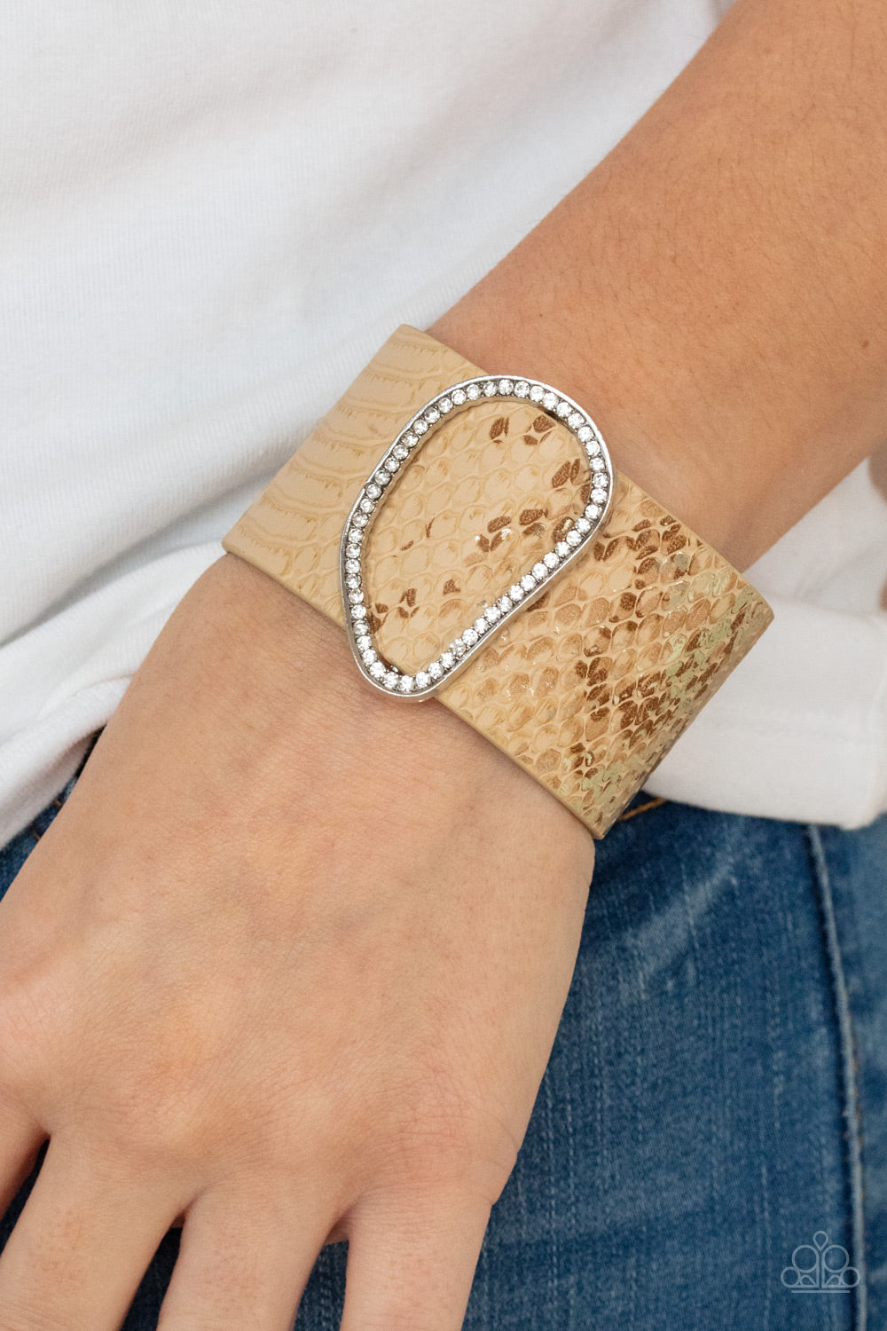 Encrusted with glassy white rhinestones, an asymmetrical silver fitting glides along a tan leather band adorned in a golden metallic python print for a wild look