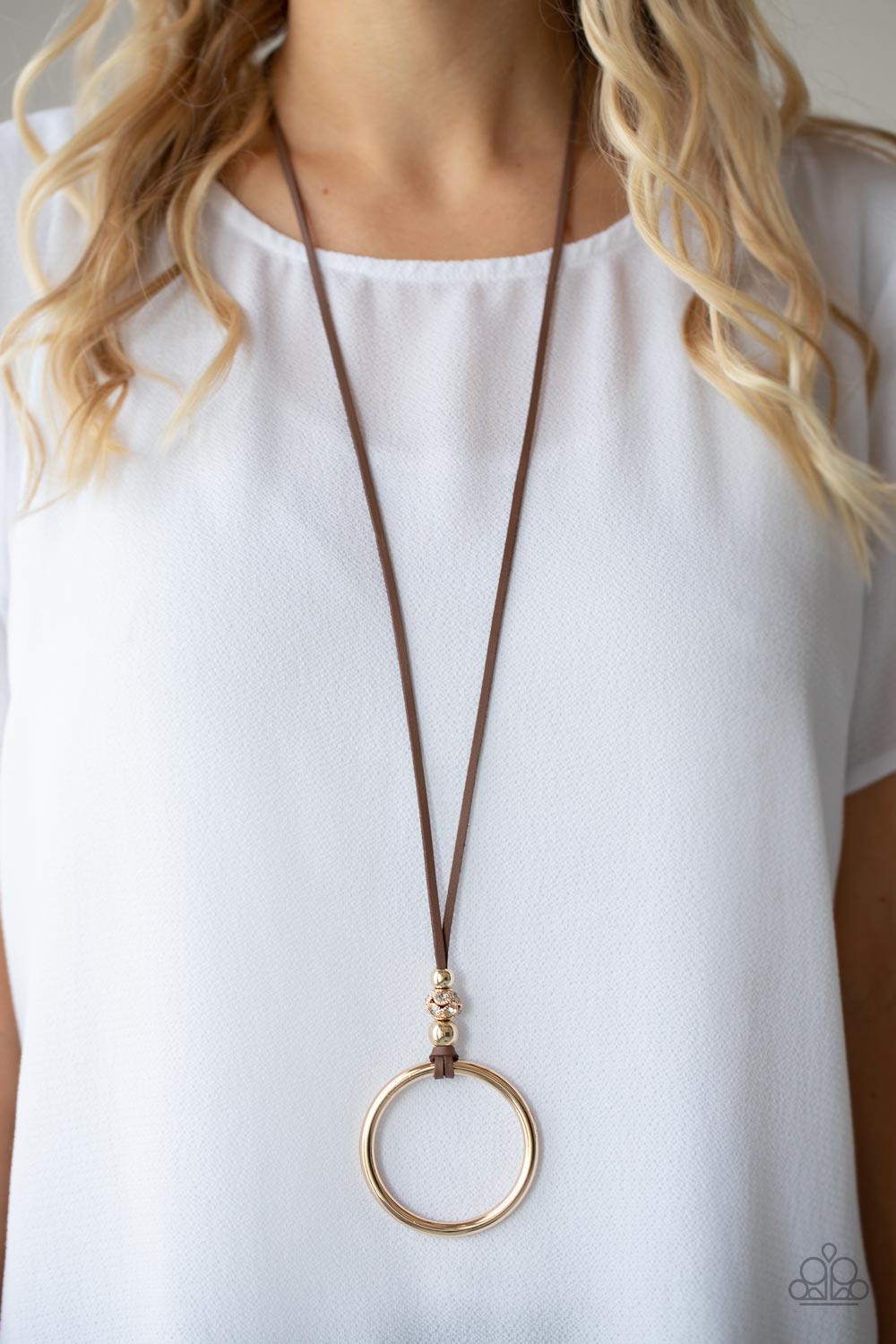 Infused with pair of classic gold beads and a white rhinestone encrusted accent, lengthened brown leather cording knots around a bold gold hoop 