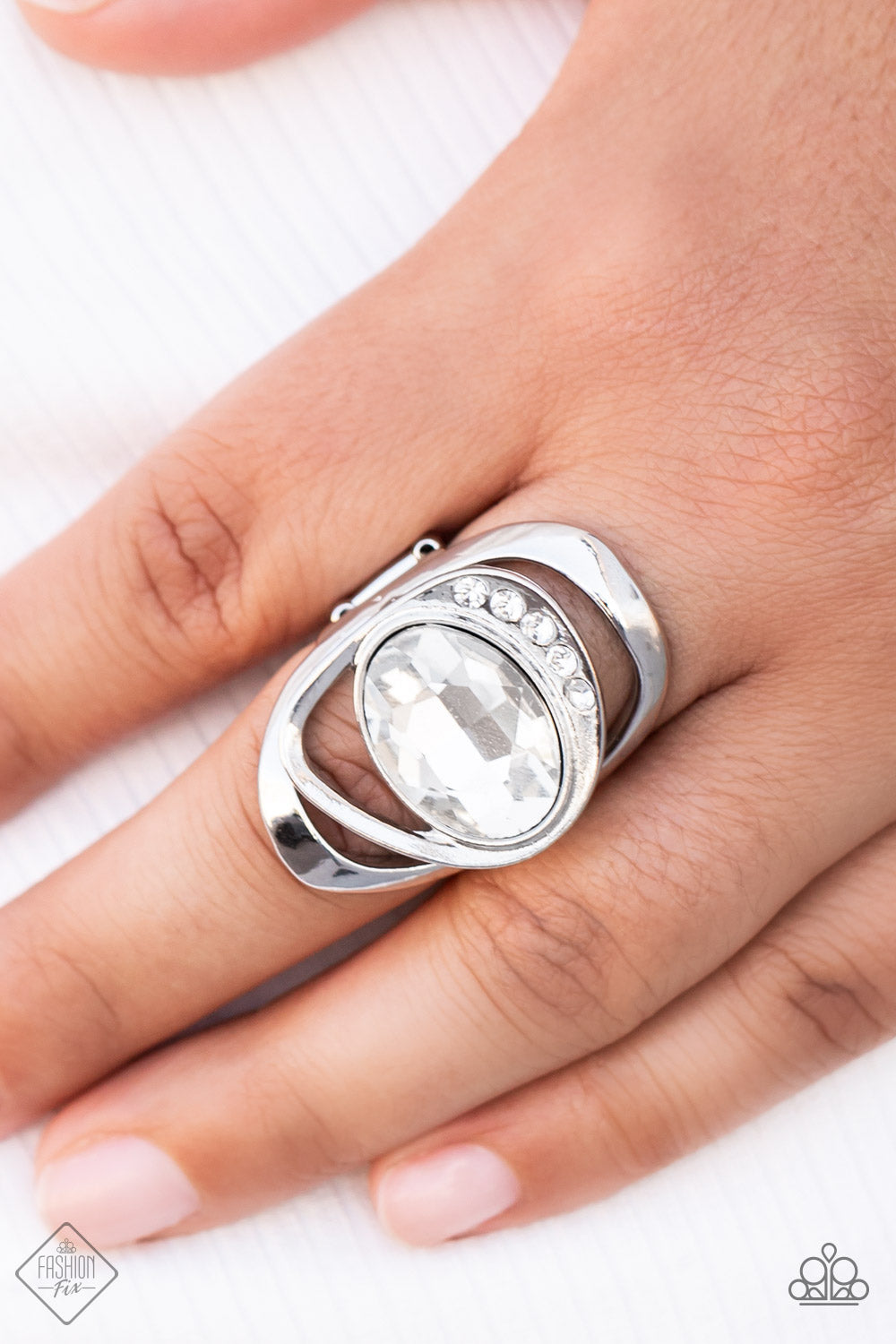 oversized white oval gem is tilted on its side and nestled into a sleek silver frame. A row of dainty white rhinestones runs along the edge
